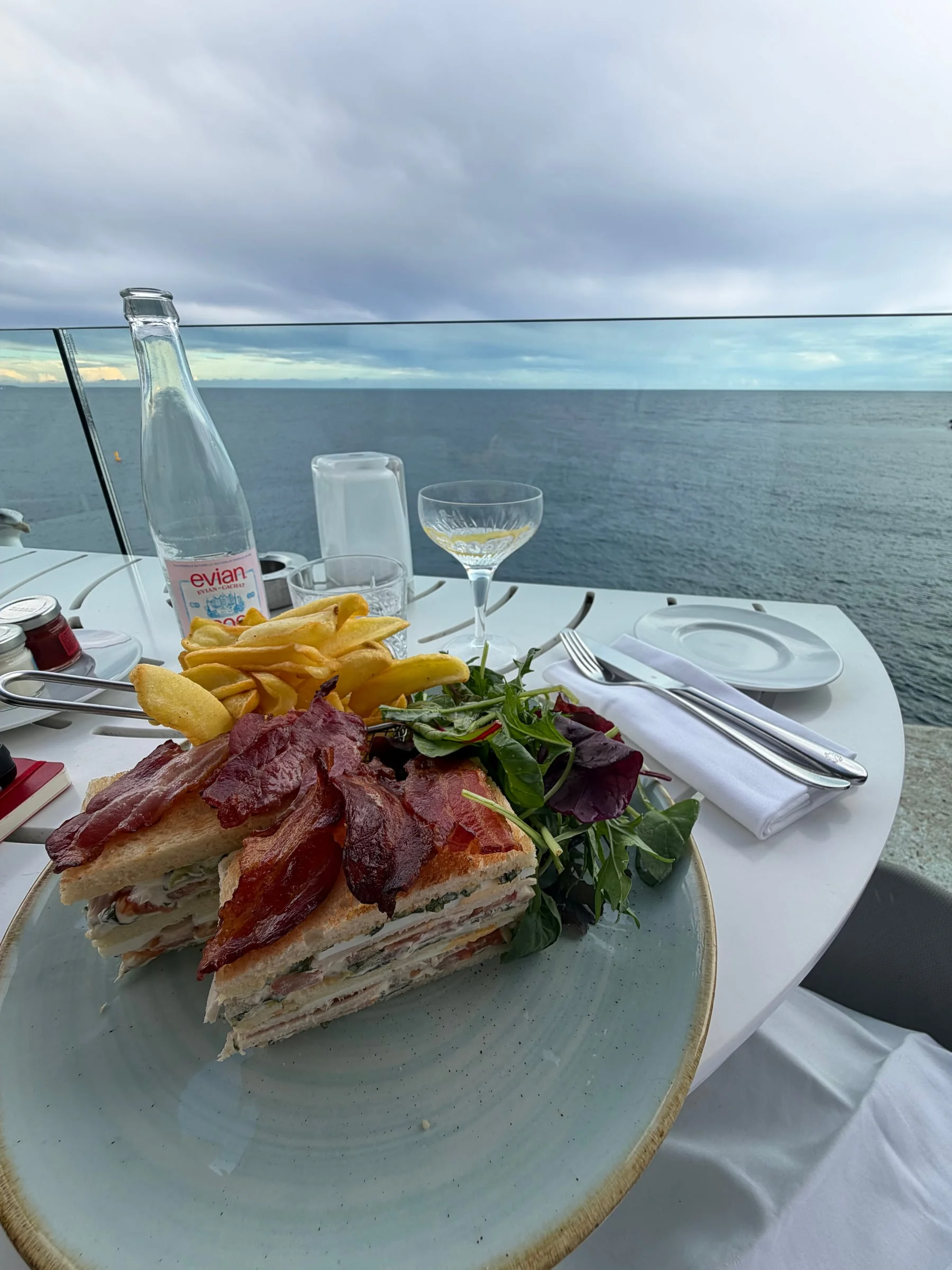 A tall club sandwich cut in half with fries and salad, on a terrace table above the Mediterranean